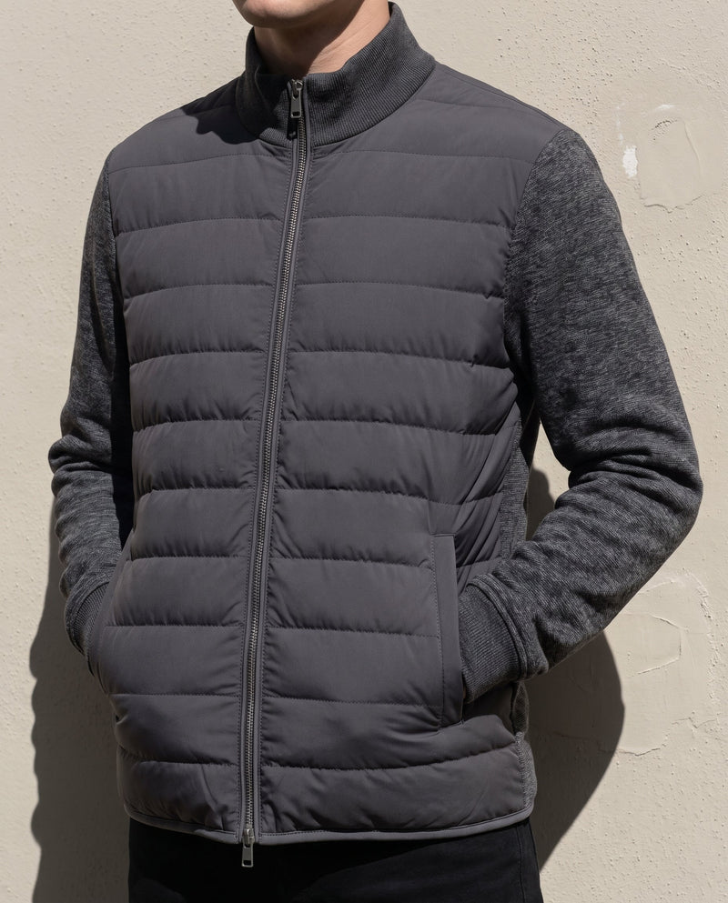 Bellardi Hybrid Puffer Jacket in Dark Grey, worn closed with hands in the side pockets, showing the quilted front panel and knit sleeves against a painted plaster wall.