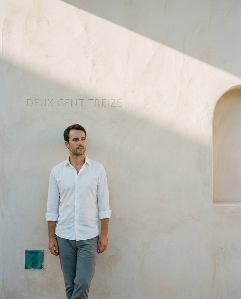 A man in a plain white shirt leans against a limewashed wall beside an arched niche, with DEUX CENT TREIZE cut small into the plaster above him.