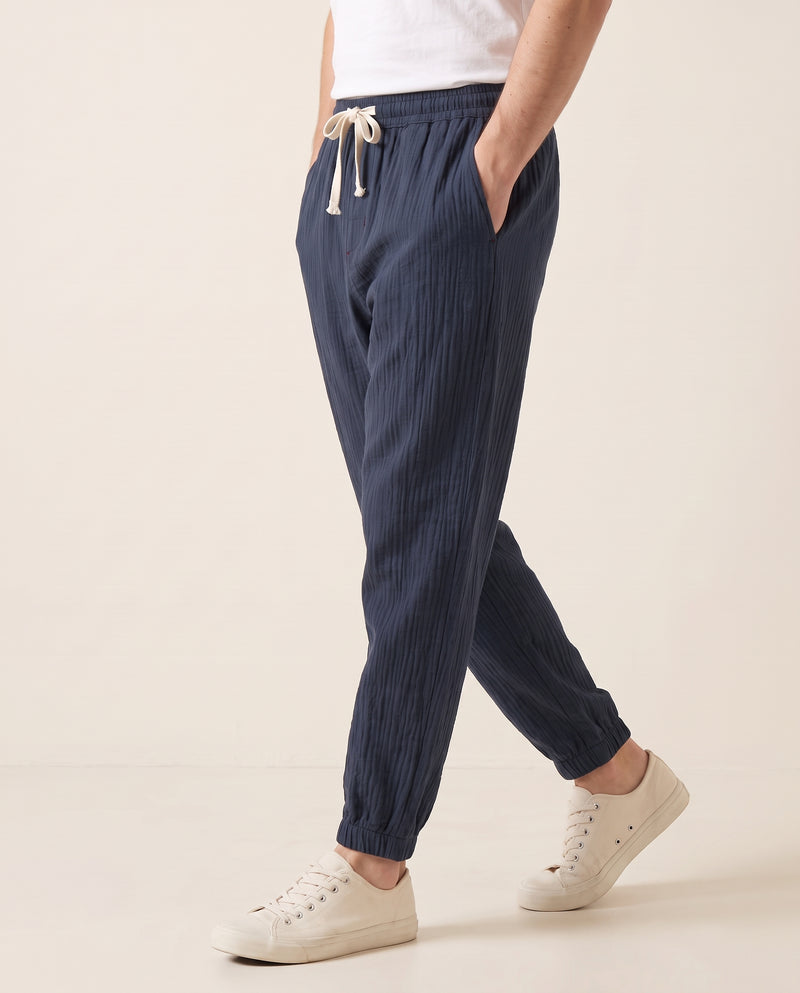 Homme en marche portant un pantalon confort bleu marine, tissage froissé, bas de jambe élastiqué, vue de profil sur fond studio blanc cassé.