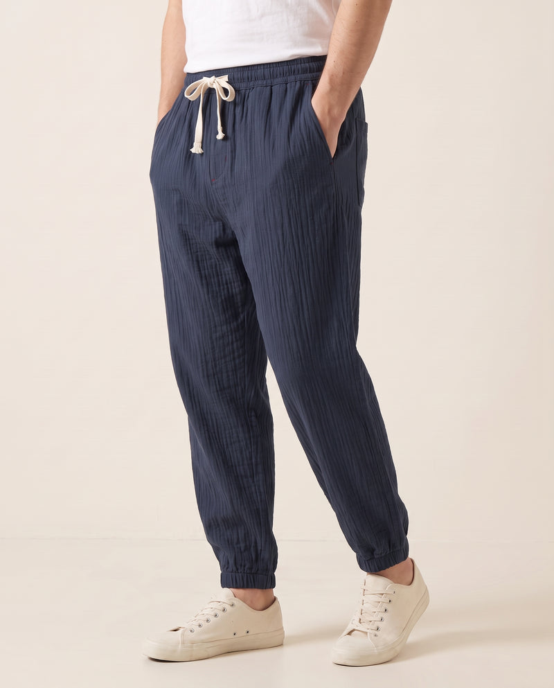 Homme portant un pantalon confort bleu marine, vue de trois quarts montrant la jambe fuselée et le cordon à la taille, tissage froissé, fond studio blanc cassé.