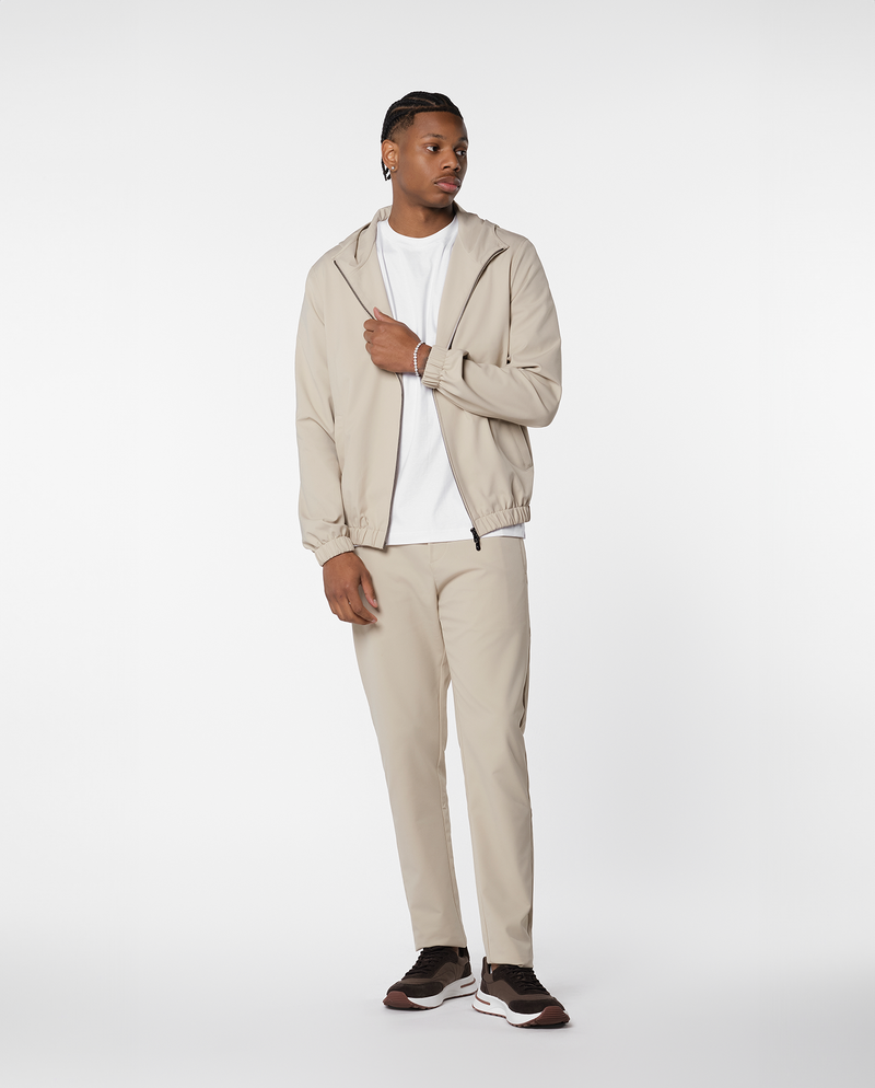 Homme portant une veste coupe-vent à capuche beige ouverte, vue de trois quarts en pied