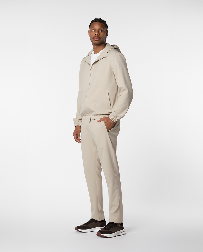 Homme portant une veste coupe-vent à capuche beige et le pantalon assorti, vue de profil en pied