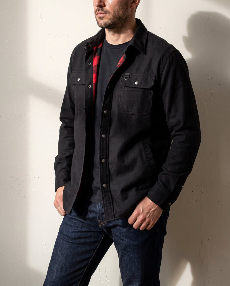 Homme portant une surchemise noire doublée de flanelle, ouverte au col pour révéler la doublure à carreaux buffalo rouge et noir, mains dans les poches, devant un mur en plâtre