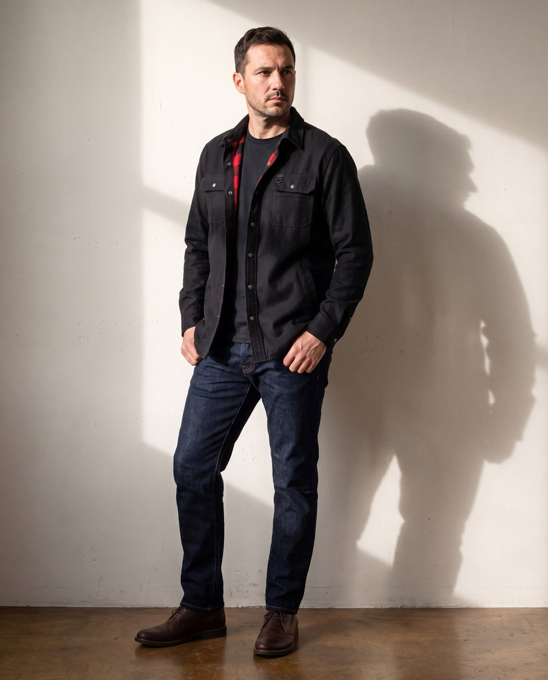 Photo en pied d'un homme portant la surchemise noire doublée de flanelle sur un t-shirt marine et un jean brut, chaussures en cuir marron, devant un mur en plâtre en lumière naturelle