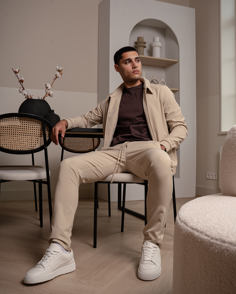 Homme en veste zippée beige et pantalon assorti, assis sur une chaise dans un intérieur aux tons chauds