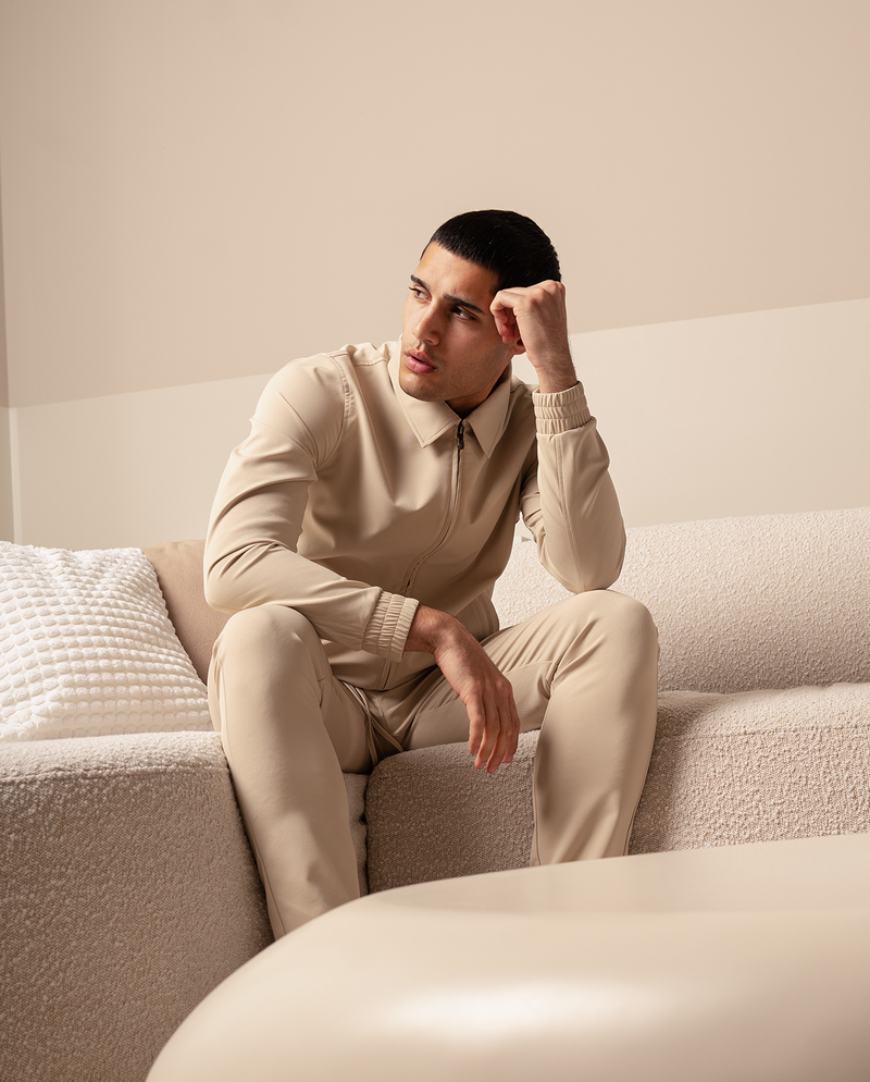Homme en veste zippée beige et pantalon assorti, assis sur un canapé crème